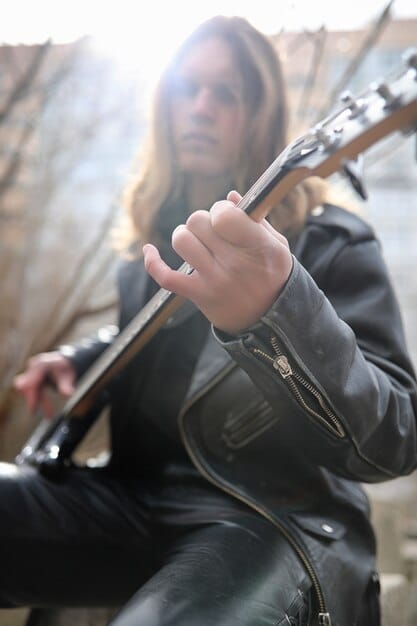 A close-up shot of a young female rock guitarist shredding on her electric guitar during a live performance. Her face is focused and intense, and her fingers are flying across the fretboard. The background is blurred, highlighting her performance.